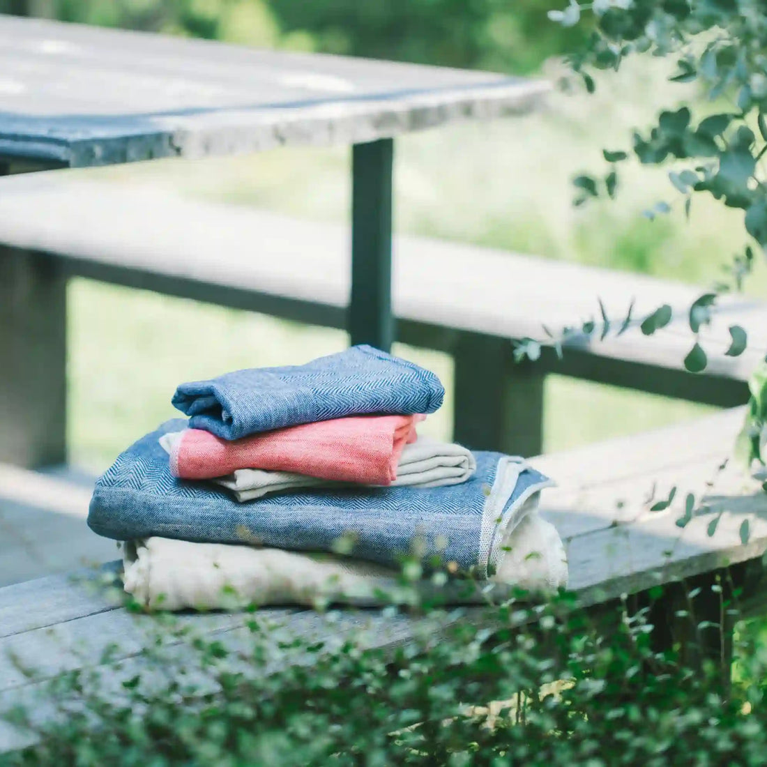 A stack of folded folded towels or cloths in blue, coral pink, and cream, placed on a wooden bench in an outdoor garden setting.