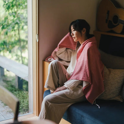A woman sits on a blue sofa in a cozy room, wrapped in a UMI red bath towel. Acoustic guitars are hanging on the wall above her, and a large window on the left shows green foliage outside.