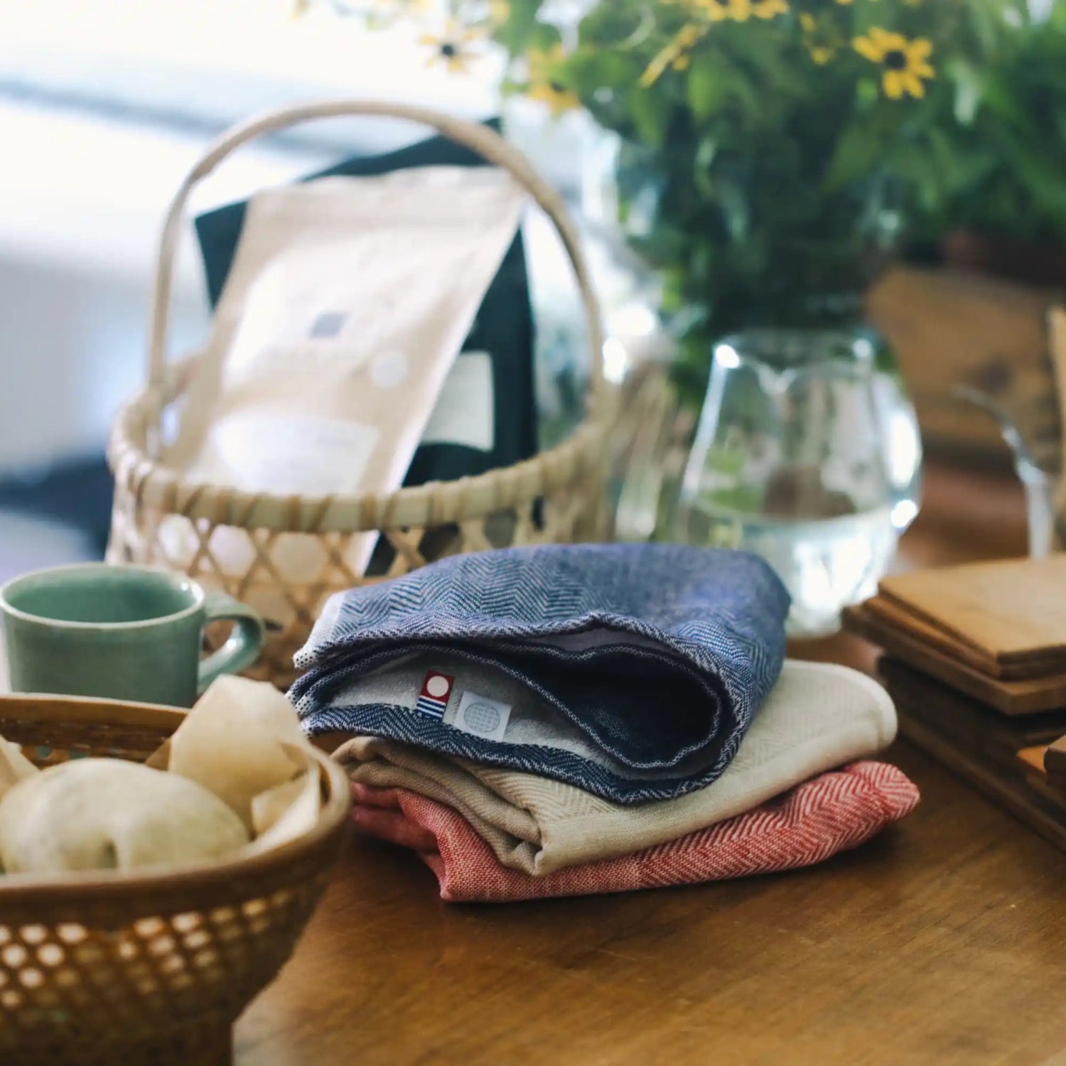 A styled tabletop scene with folded fabric cloths in blue, cream, and pink, alongside a woven basket, a small green cup, wooden coasters, and soft-focus background decor including flowers and glassware.