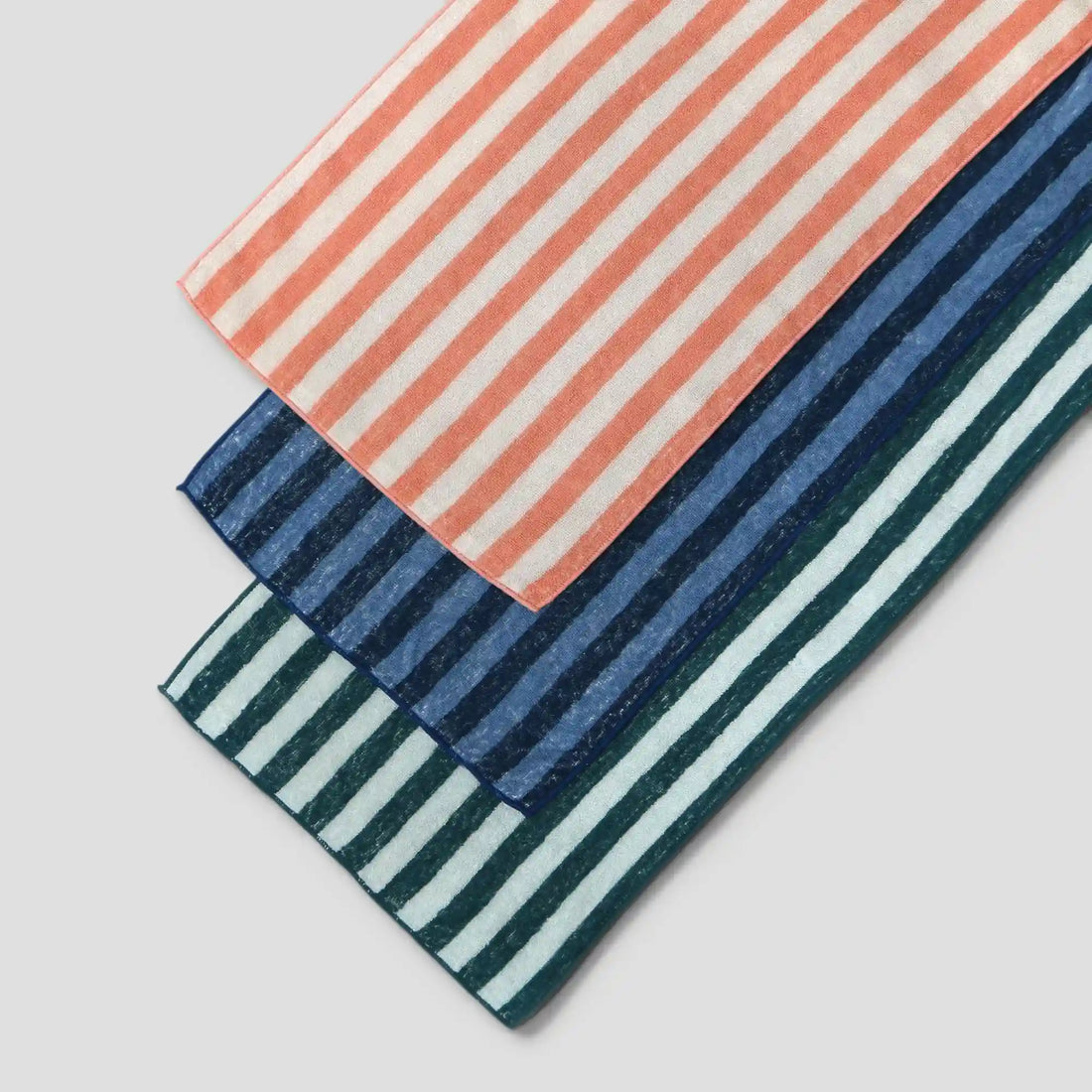 Three striped Imabari towels, one with pink and white stripes , with navy and blue stripes, green and white stripes ,on a light gray background.