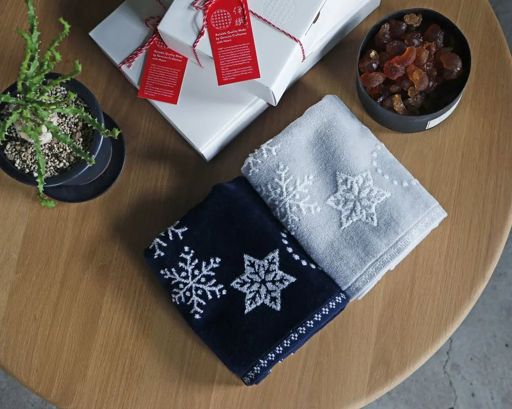 Two folded towels with snowflake patterns on a wooden table with a plant and gift boxes.